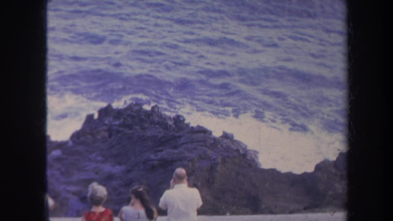 Tourists in period summer clothing observe rough surf along a Honolulu rocky shoreline in this 8mm frame.