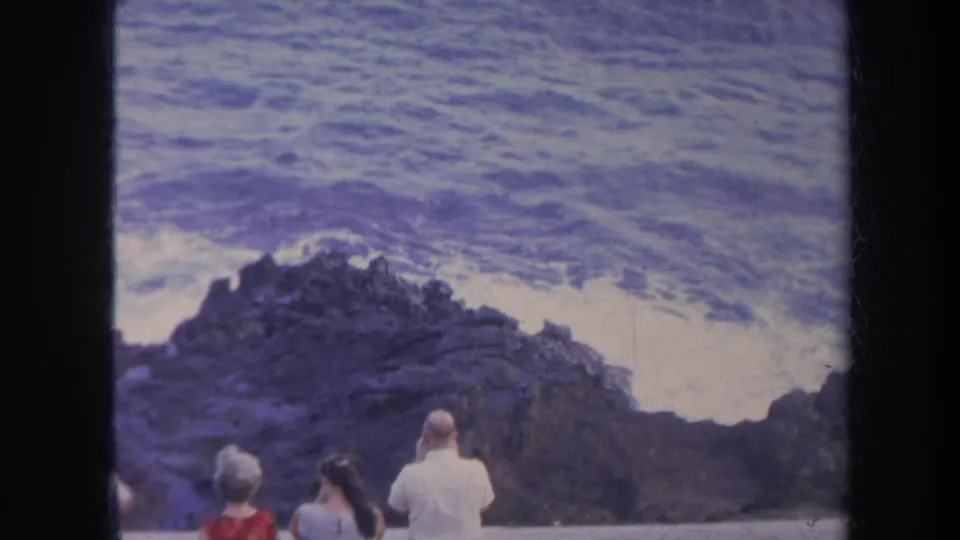 Tourists in period summer clothing observe rough surf along a Honolulu rocky shoreline in this 8mm frame.
