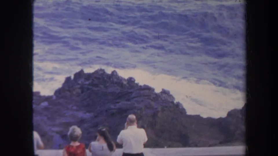 A home-movie view of visitors at a low shoreline barrier as white surf breaks against dark Honolulu rocks.