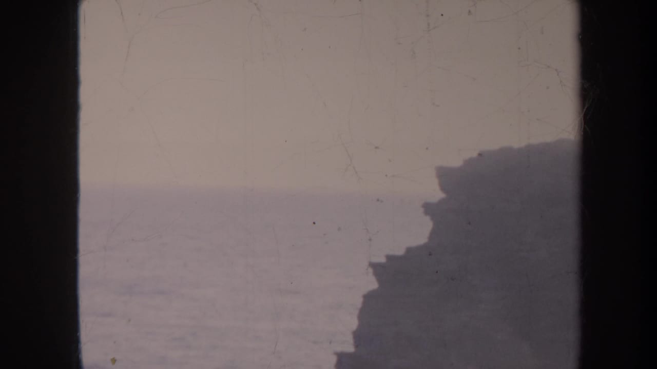 1967 Honolulu 8mm frame showing a muted ocean horizon and dark coastal cliff, with dust and scratches visible on the film.