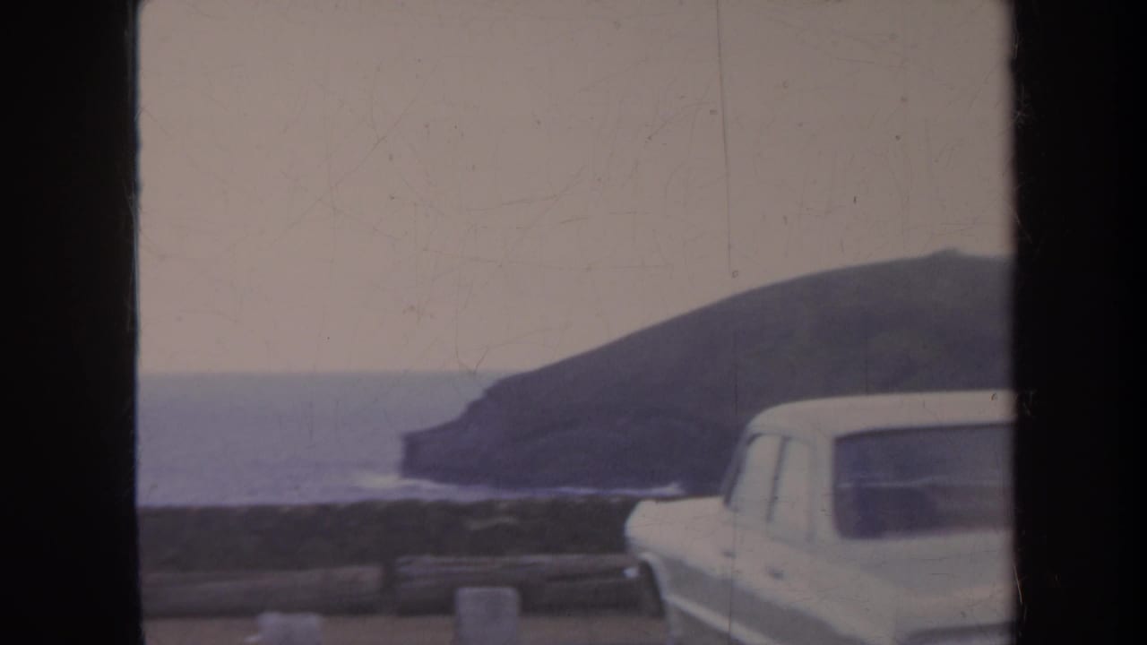 Honolulu home-movie frame from 1967 showing a white station wagon by a roadside barrier, with ocean and dark coastal headland behind it.