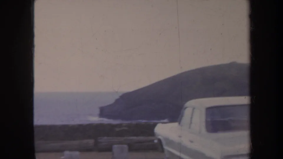 Honolulu home-movie frame from 1967 showing a white station wagon by a roadside barrier, with ocean and dark coastal headland behind it.