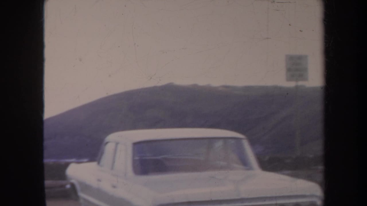 1967 Honolulu-area home-movie frame showing a white mid-century sedan near a misty coastal landscape, with dark frame bands from the scan.