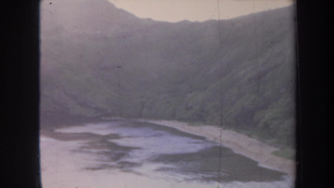1967 Honolulu 8mm frame showing a small beach, muted green hills, and calm shoreline water under a pale sky.