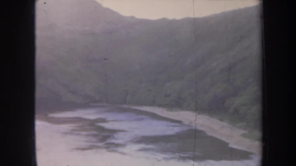 1967 Honolulu 8mm frame showing a small beach, muted green hills, and calm shoreline water under a pale sky.