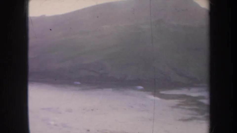 1967 Honolulu home-movie frame of a broad hillside coastline, still water, and light surf marks along the shore.