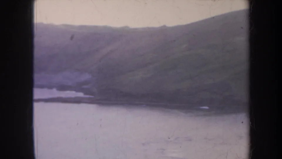 1967 Honolulu 8mm frame showing a long green hill above quiet coastal water and a narrow shoreline in soft focus.