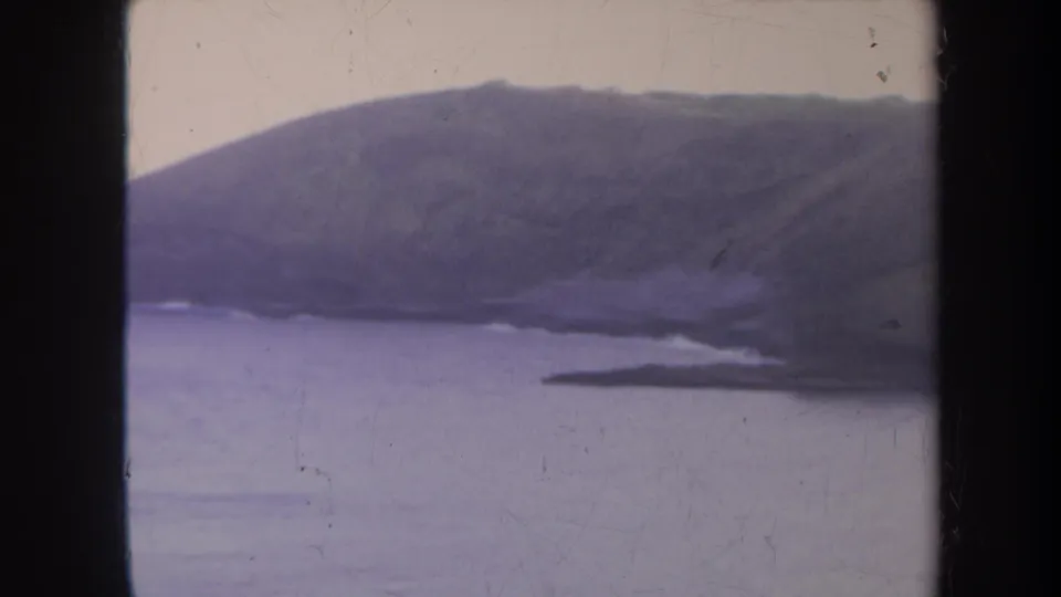 1967 Honolulu home-movie frame with a rounded green hill, calm water, pale sky, and a small point of shoreline.