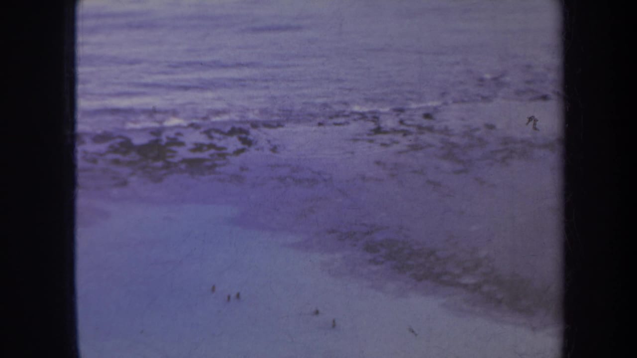 1967 Honolulu-area beach view with purple-tinged surf, rocky shallows, and distant people near the shoreline.
