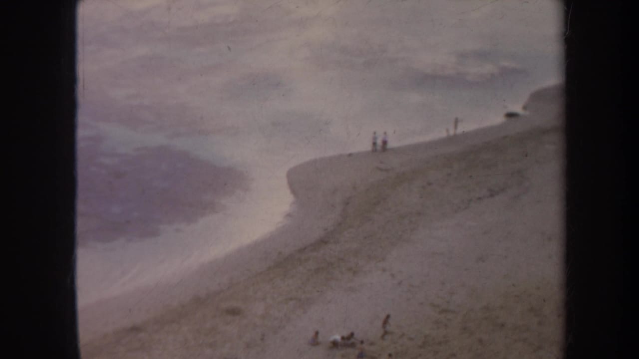 1967 Honolulu 8mm frame showing a sandy shoreline, calm water, and small distant figures near the top of the slope.