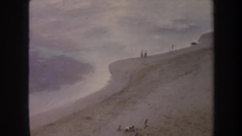 1967 Honolulu 8mm frame showing a sandy shoreline, calm water, and small distant figures near the top of the slope.