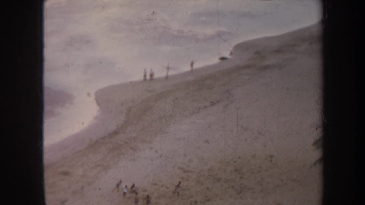 Sandy hill beside water with several distant figures and dark vertical film borders.