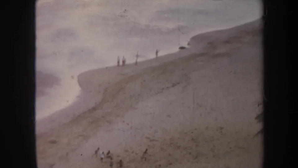 1967 Honolulu home-movie frame of a sandy coastal slope, calm inlet water, and tiny distant people near the ridge.