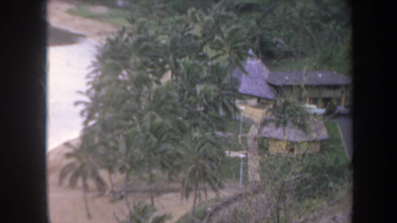 1967 Honolulu 8mm frame showing a tropical compound with palm trees, thatched roofs, sandy beach, and calm water beyond a dark window frame.