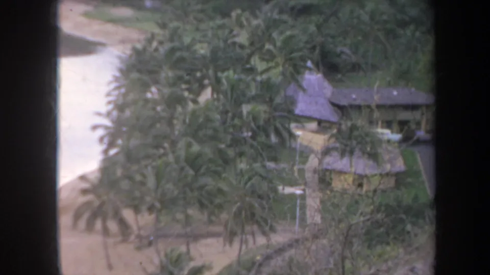 1967 Honolulu 8mm frame showing a tropical compound with palm trees, thatched roofs, sandy beach, and calm water beyond a dark window frame.