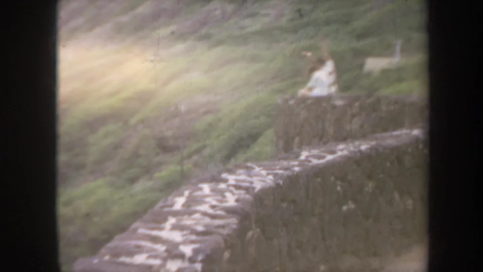 1967 Honolulu 8mm frame showing a stone wall and green hillside, with two distant people seated near the overlook area.