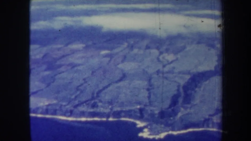 1967 Hawaii aerial home-movie view showing rugged volcanic terrain, pale ridges, and a dark coastal edge.
