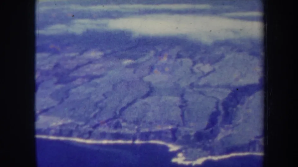 Blue-tinted 8mm aerial view of Hawaii landscape, with volcanic ridges and a shoreline visible near the lower frame.
