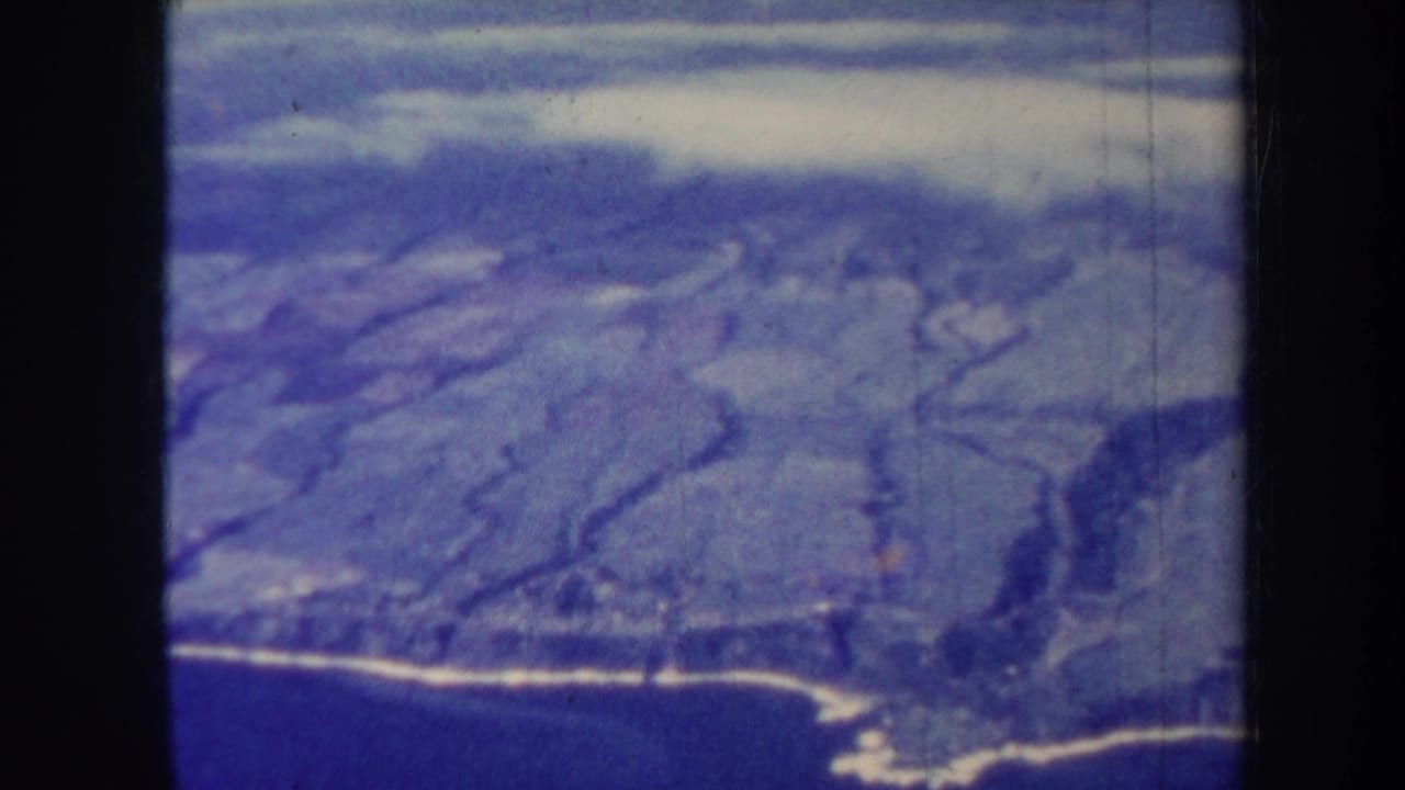 1967 Honolulu home-movie frame showing a soft blue aerial view of shoreline, hills, and ocean from an airplane-window perspective.
