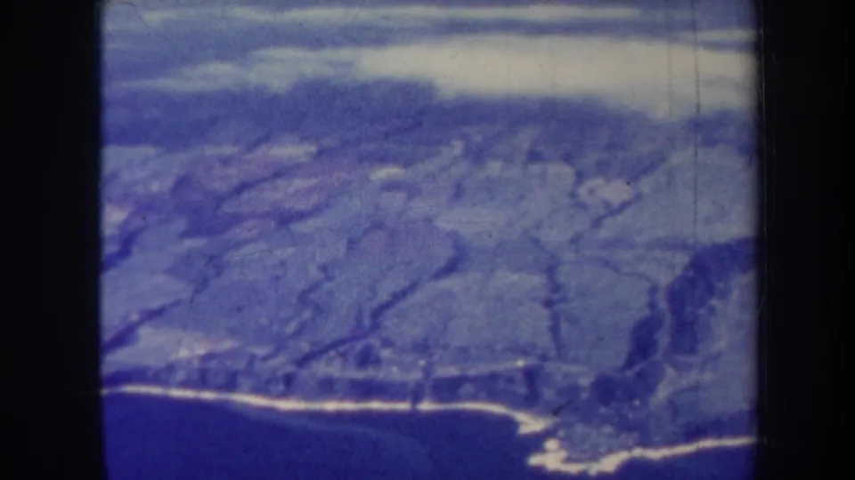 1967 Honolulu aerial frame with coastline, hillside terrain, and ocean rendered in blue-tinted 8mm home-movie color.
