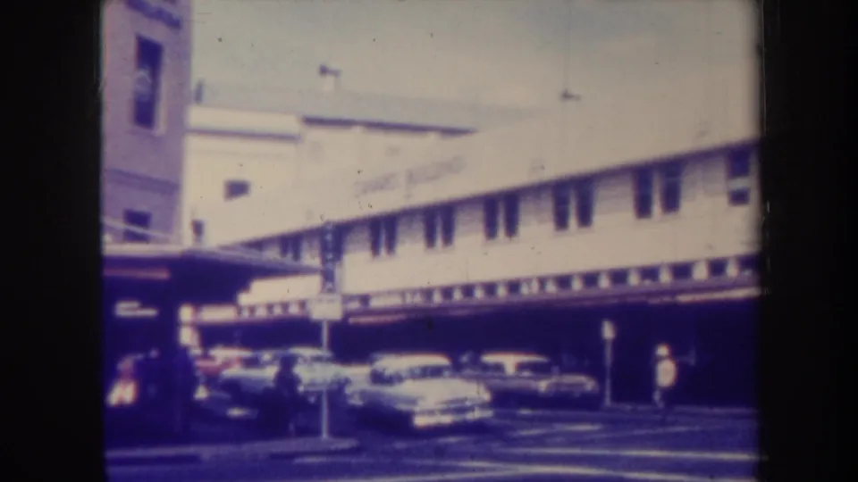 1967 Honolulu street frame showing a two-story commercial building, older cars at the curb, street markings, and small pedestrian figures.
