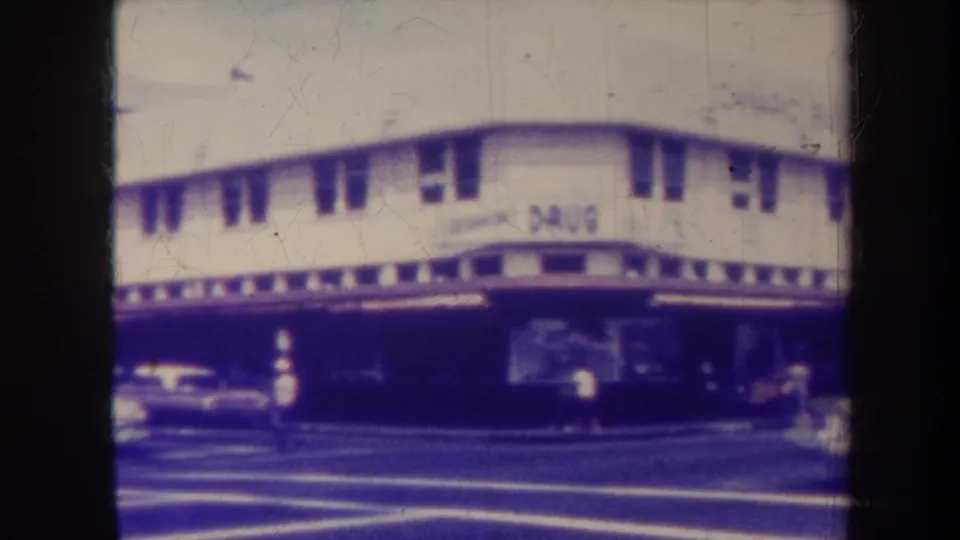 1967 Honolulu street scene with a visible drugstore sign on a two-story corner building, older cars, crosswalk markings, and pedestrians.
