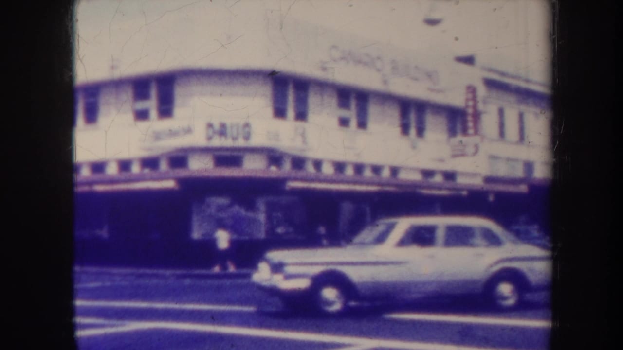 1967 Honolulu street view with a white sedan moving past storefronts and a building sign.