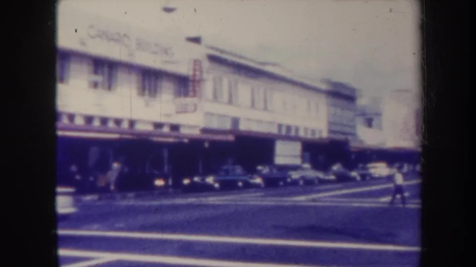 1967 Honolulu commercial block with classic cars, storefront awnings, and a pedestrian near the crossing.