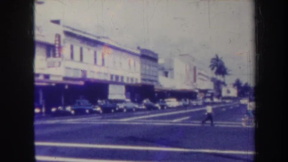 1967 Honolulu street scene with a pedestrian crossing in front of parked classic cars and low commercial buildings.