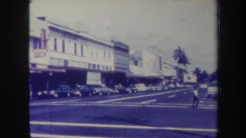 1967 Honolulu street view showing a pedestrian in the roadway, classic cars, palm trees, and a commercial block.