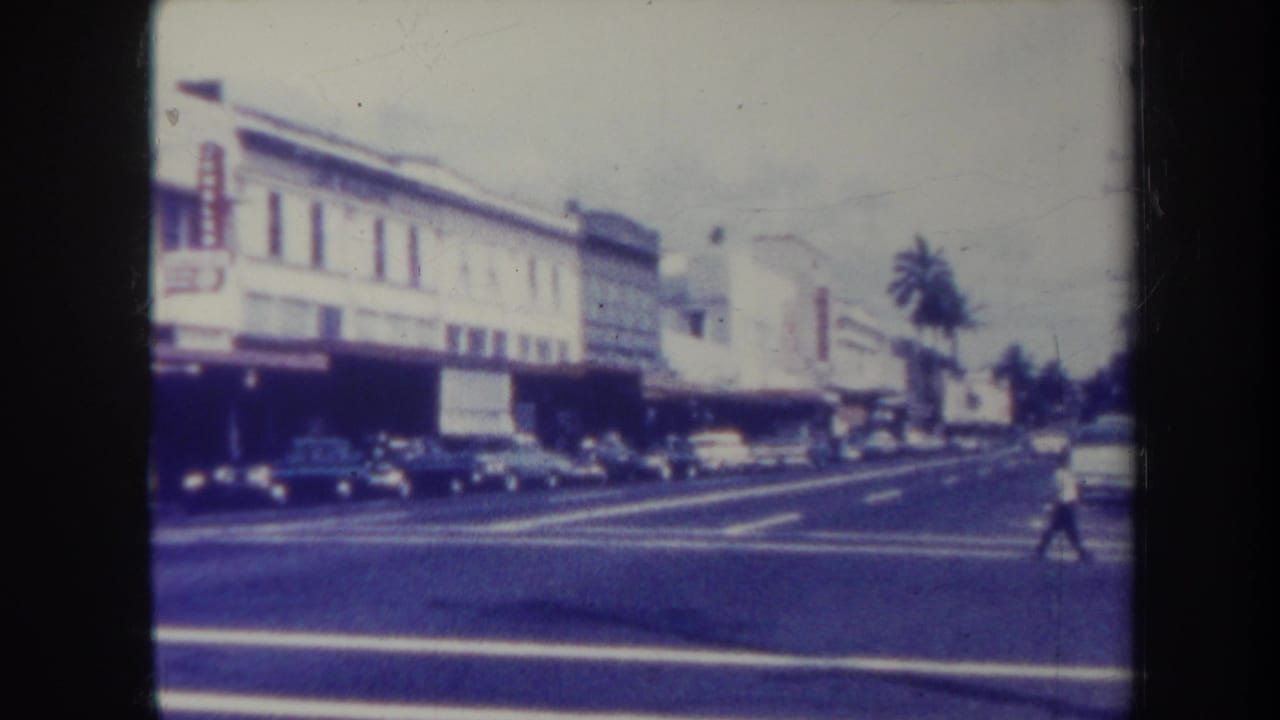1967 Honolulu street view with storefronts, parked cars, palm trees, and a pedestrian crossing in the distance.