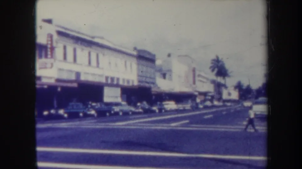 1967 Honolulu street view with storefronts, parked cars, palm trees, and a pedestrian crossing in the distance.