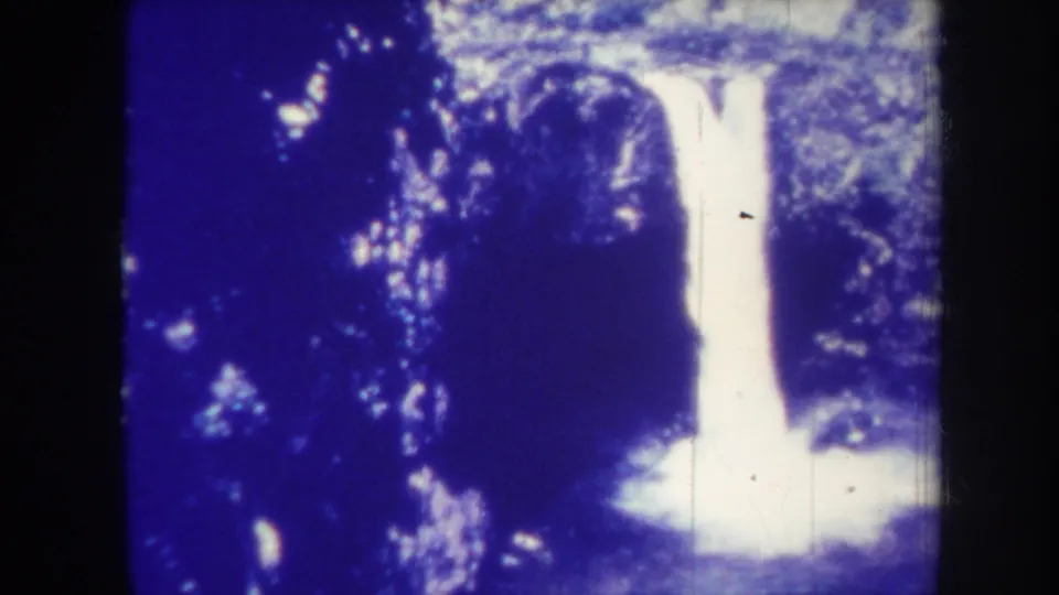 1967 Honolulu travel frame with a centered waterfall, shadowed vegetation, and overexposed white water at the base.