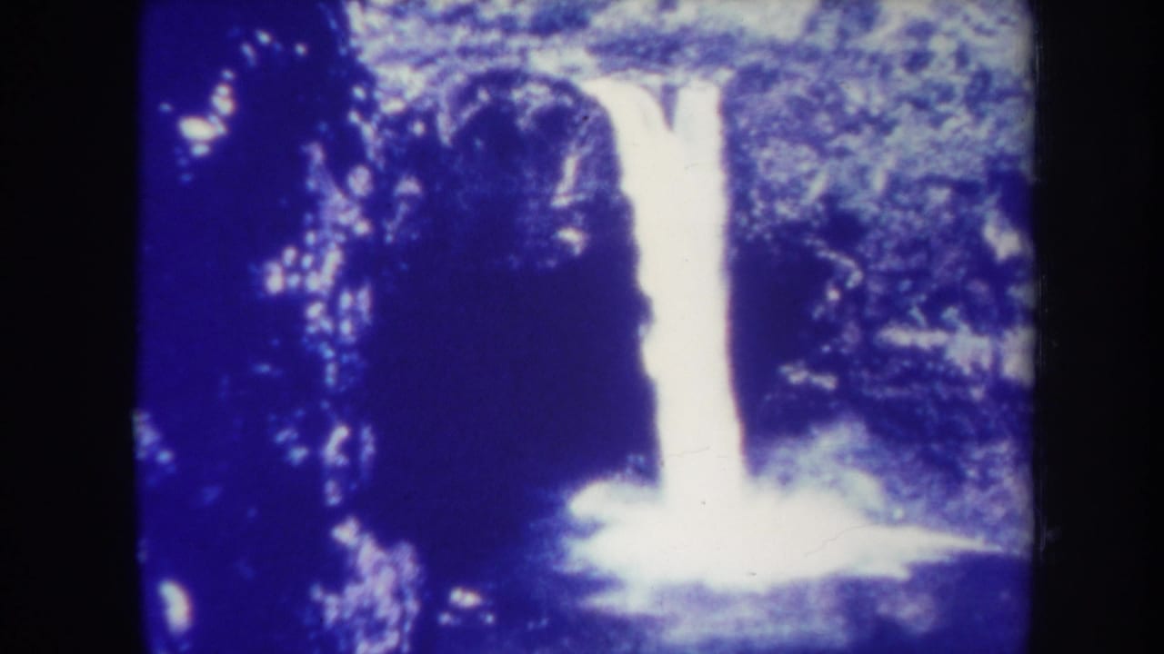 1967 Honolulu home-movie frame showing a bright waterfall against dark rock and dense tropical foliage, with blue color shift and rounded film edges.