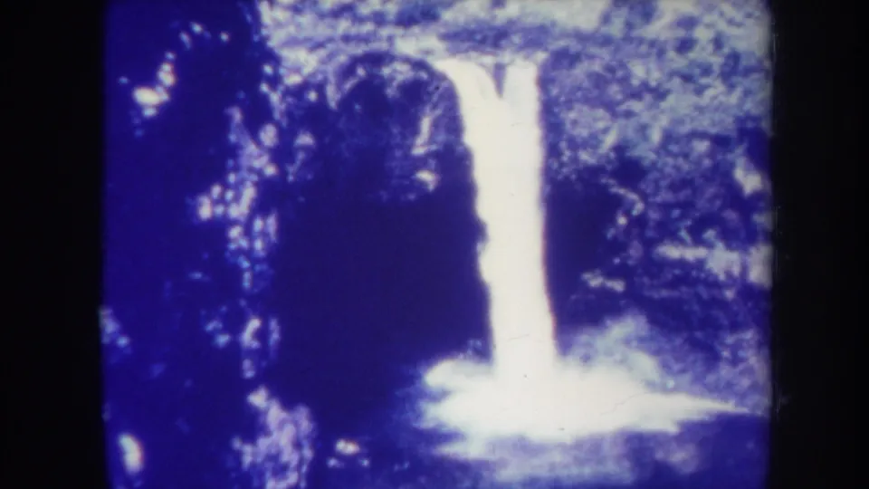 1967 Honolulu home-movie frame showing a bright waterfall against dark rock and dense tropical foliage, with blue color shift and rounded film edges.
