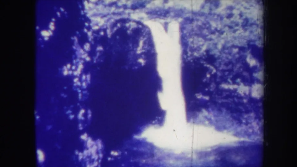 1967 Honolulu 8mm frame with a bright waterfall descending beside dark rock and dense greenery, softened by grain and exposure bloom.