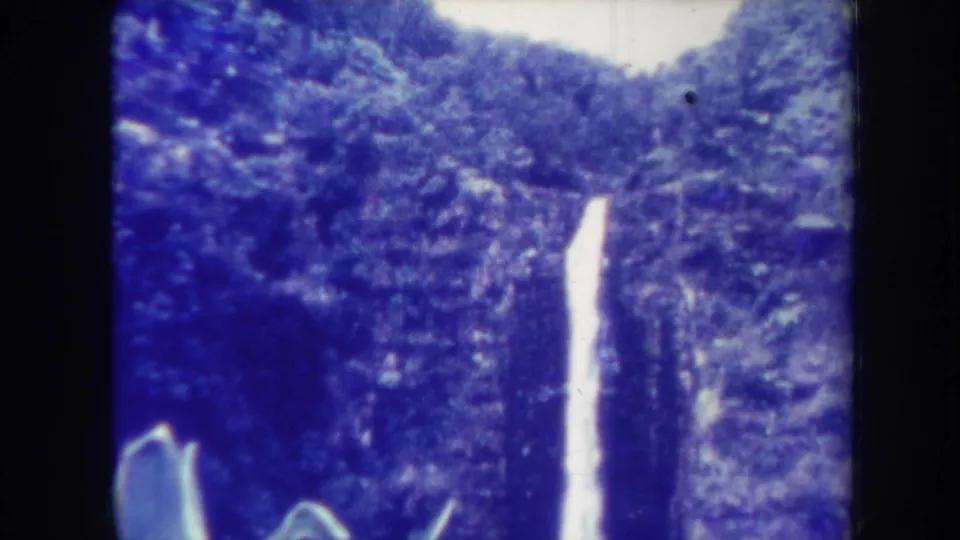 1967 Honolulu home-movie frame showing a narrower view of a waterfall dropping through dense tropical vegetation, with visible 8mm grain and gate borders.