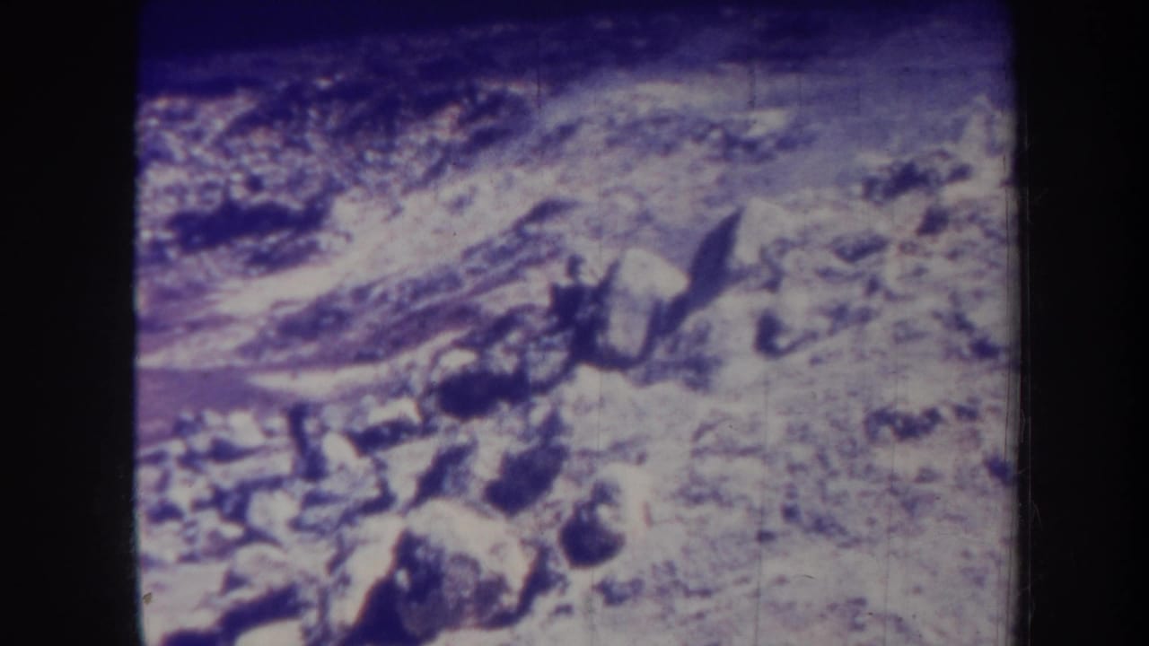 1967 Honolulu home-movie frame showing pale rocky ground, dark stones, and faint drifting smoke or haze with purple 8mm color fading.
