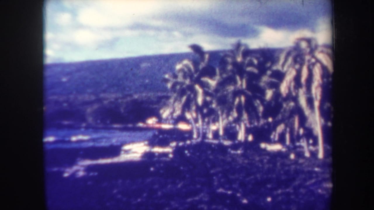 1967 Honolulu home-movie frame showing palm trees along a coastal edge, pale surf, and a dark hillside under a cloudy sky.