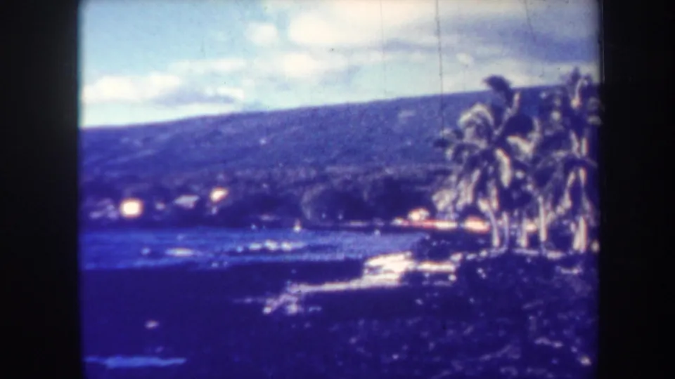 Blue-tinted 1967 Honolulu home-movie frame with shoreline palms, water, low surf, and a dark hillside in the distance.