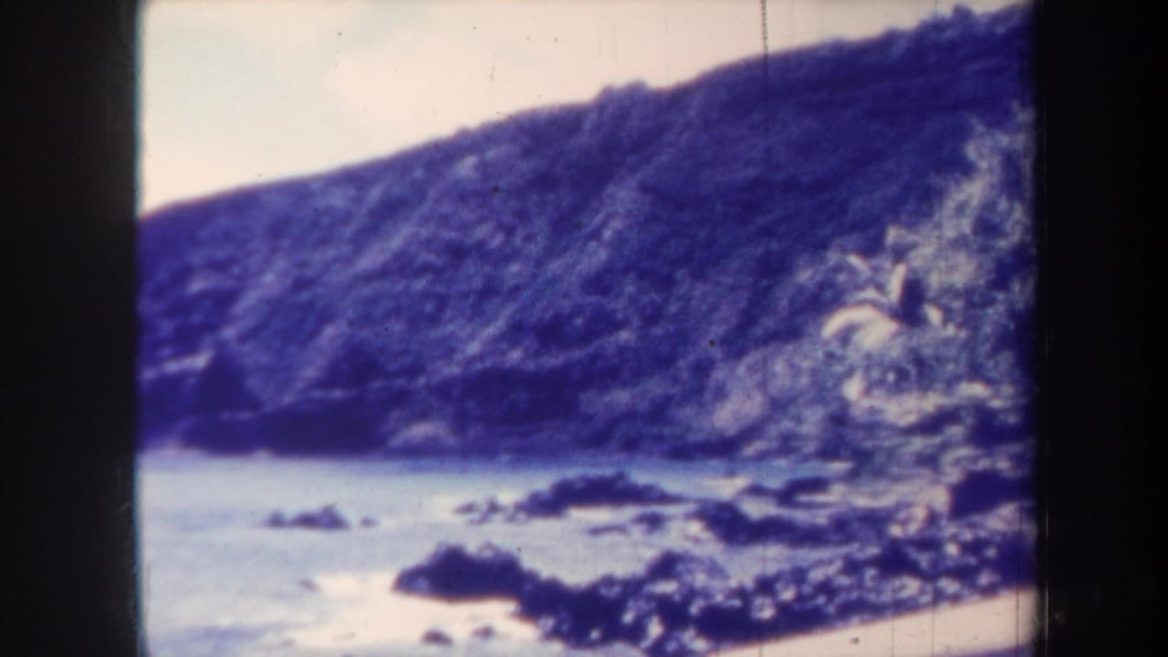1967 Honolulu home-movie frame showing dark coastal rocks, pale water, and a steep cliff under a washed sky.