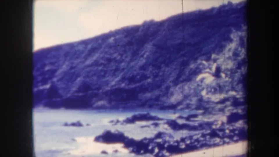 1967 Honolulu home-movie frame showing dark coastal rocks, pale water, and a steep cliff under a washed sky.