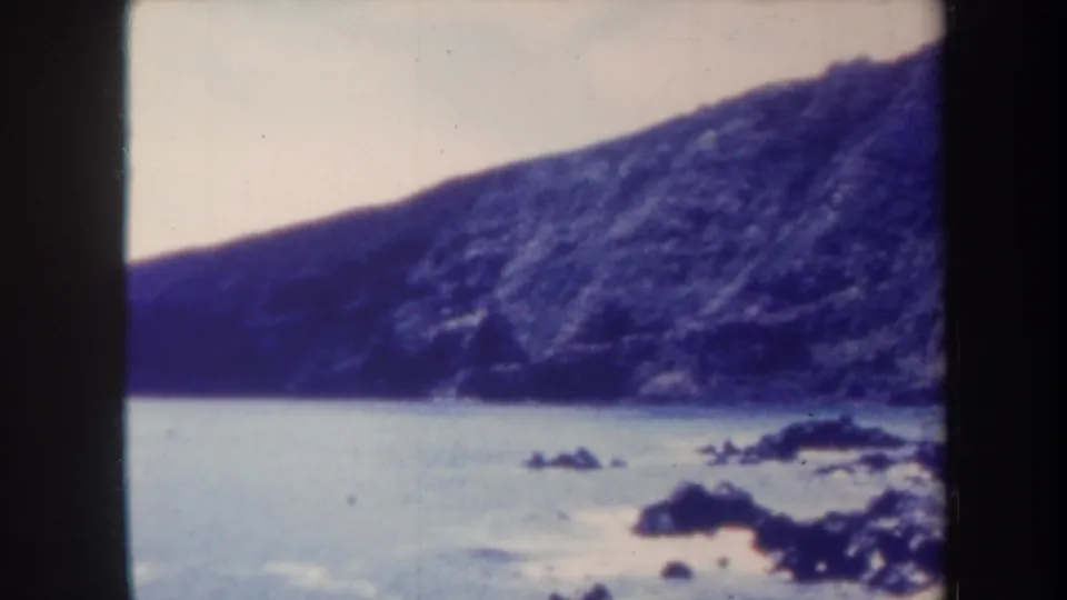 Honolulu coastal view from 1967 8mm footage, with dark rocks offshore and a broad cliff filling the background.