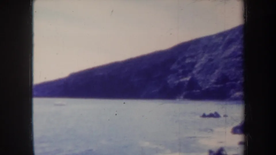 Vintage 8mm Honolulu coastline frame with calm water, a dark sloping cliff, and small rocks near the lower edge.