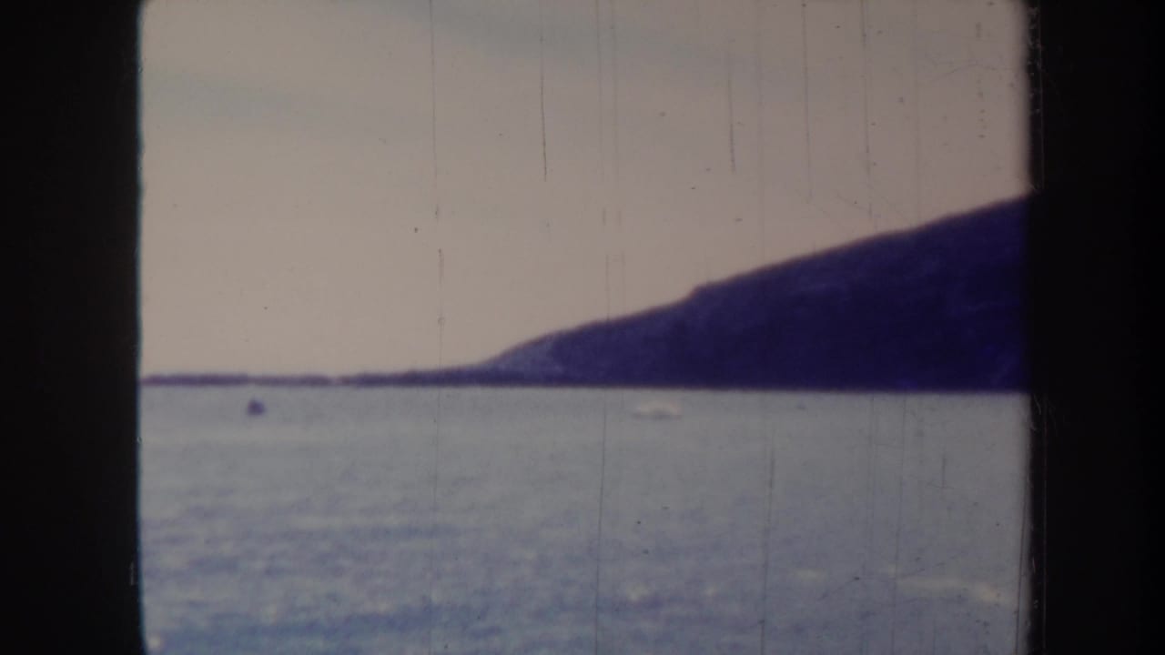 Distant headland across calm water under a pale sky, framed by black film edges.