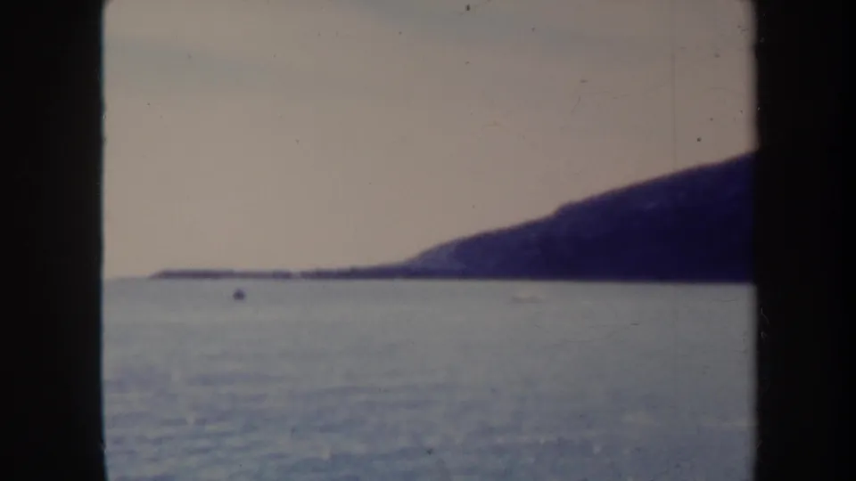1967 Honolulu-area 8mm travel footage frame with blue water, low distant shore, and a dark sloping landform under a pale sky.