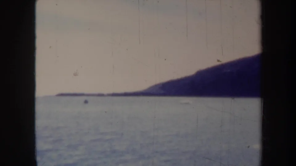 1967 Honolulu-area coastal frame showing calm water, a dark sloping shoreline, pale sky, and visible scratches from the 8mm source.