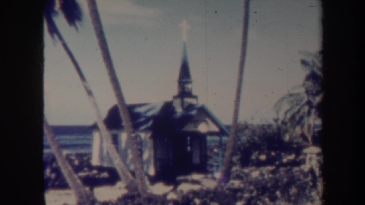 Small steepled wooden church near palms with ocean behind it.