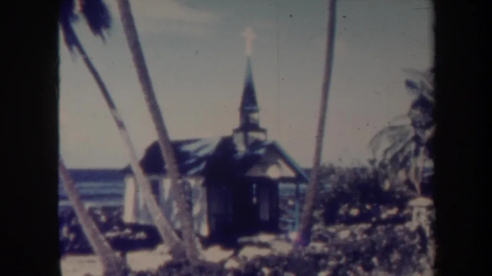 1967 Honolulu-area 8mm frame of a small wooden church with steeple and cross, framed by leaning palm trunks and ocean water.