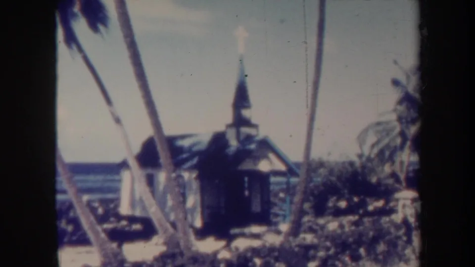 1967 Honolulu-area 8mm frame holding on the small beachside church, with palms crossing the foreground and ocean visible behind.